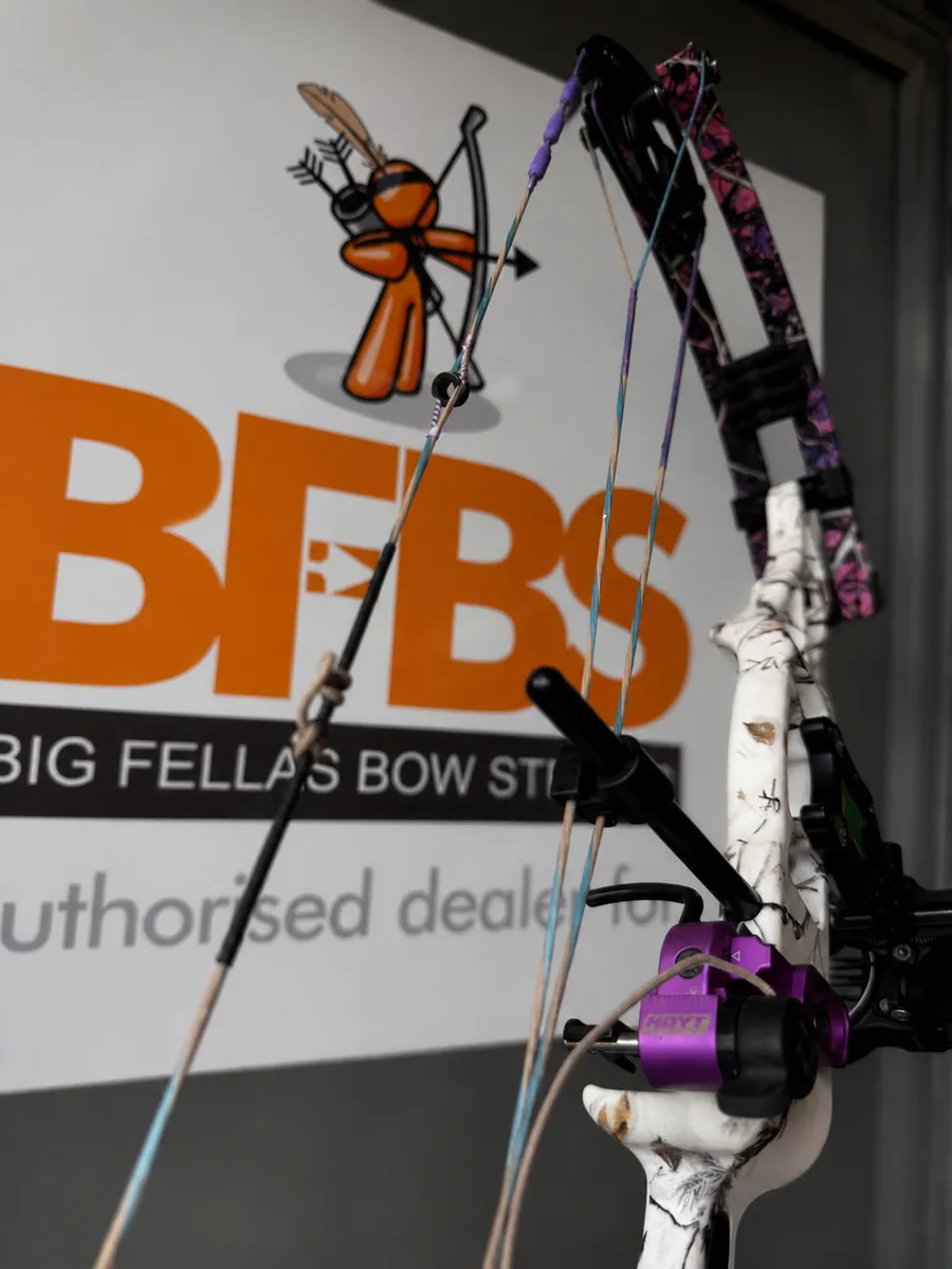 A Big Fella's Bow Strings authorised dealer sign on the wall behind a compound bow strung with purple and tan strings.
