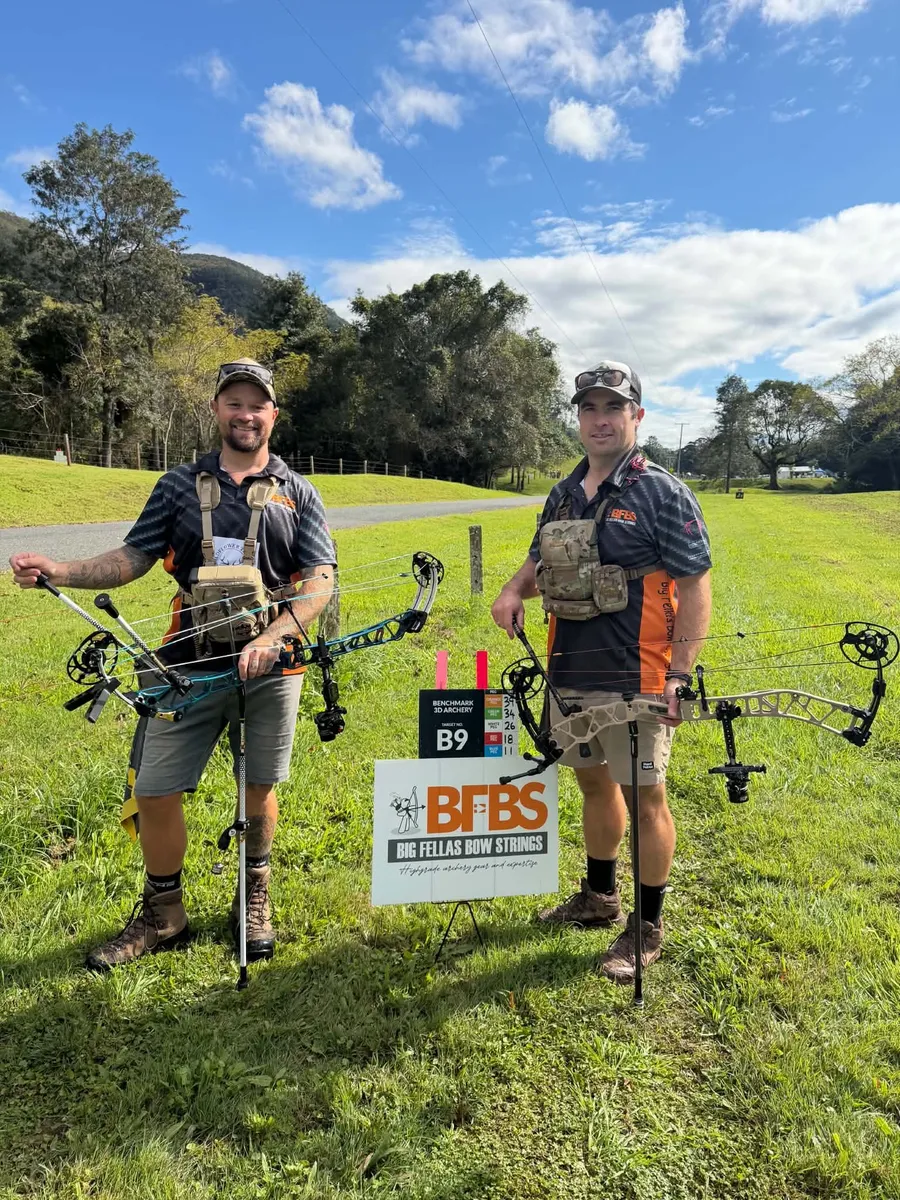 Two archers standing either side of a Big Fella's Bow Strings sign at a 3D target peg.