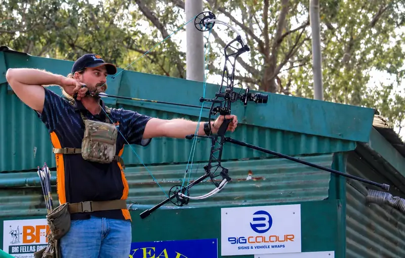 An archer at full draw at a club range with the Big Fella's banner on the shed behind.