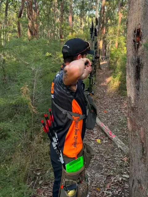 An archer at full draw on a bush slope, aiming uphill between the trees.