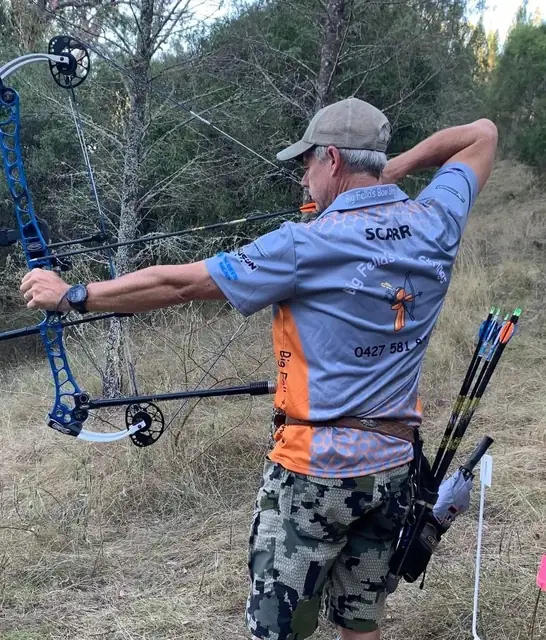 An archer at full draw in open scrub with a hip quiver of arrows.