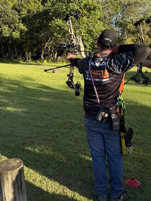 An archer at full draw on the range in a team shirt, seen from behind.
