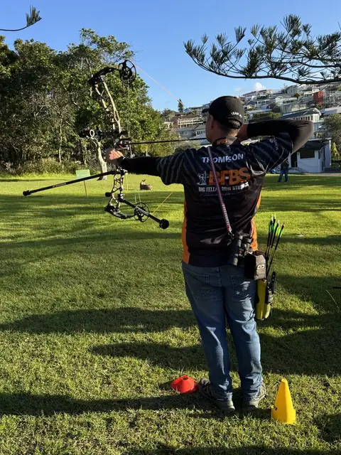 An archer at full draw on the practice lawn with marker cones underfoot.