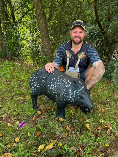 An archer kneeling behind a life-size 3D pig target on a bush course.