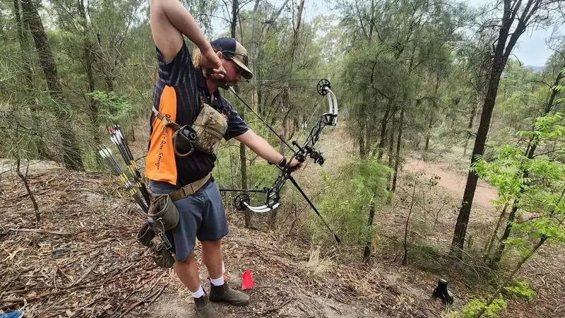 An archer in an orange team shirt at full draw on a bush course.