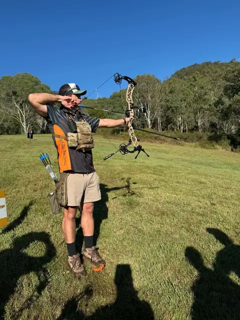 An archer at full draw on the grass with a long afternoon shadow.