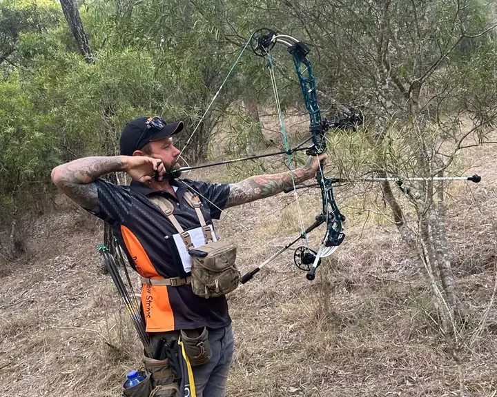 An archer at full draw on a bush course, aiming through the trees.