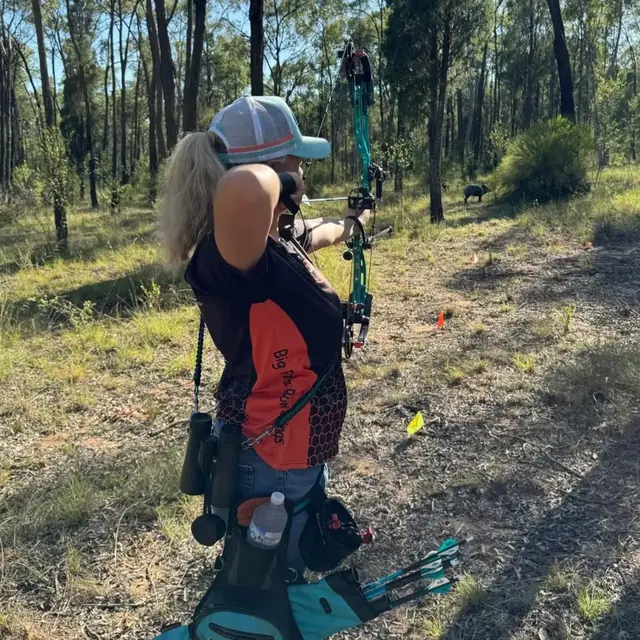 An archer aiming a teal and black compound bow across open bush.