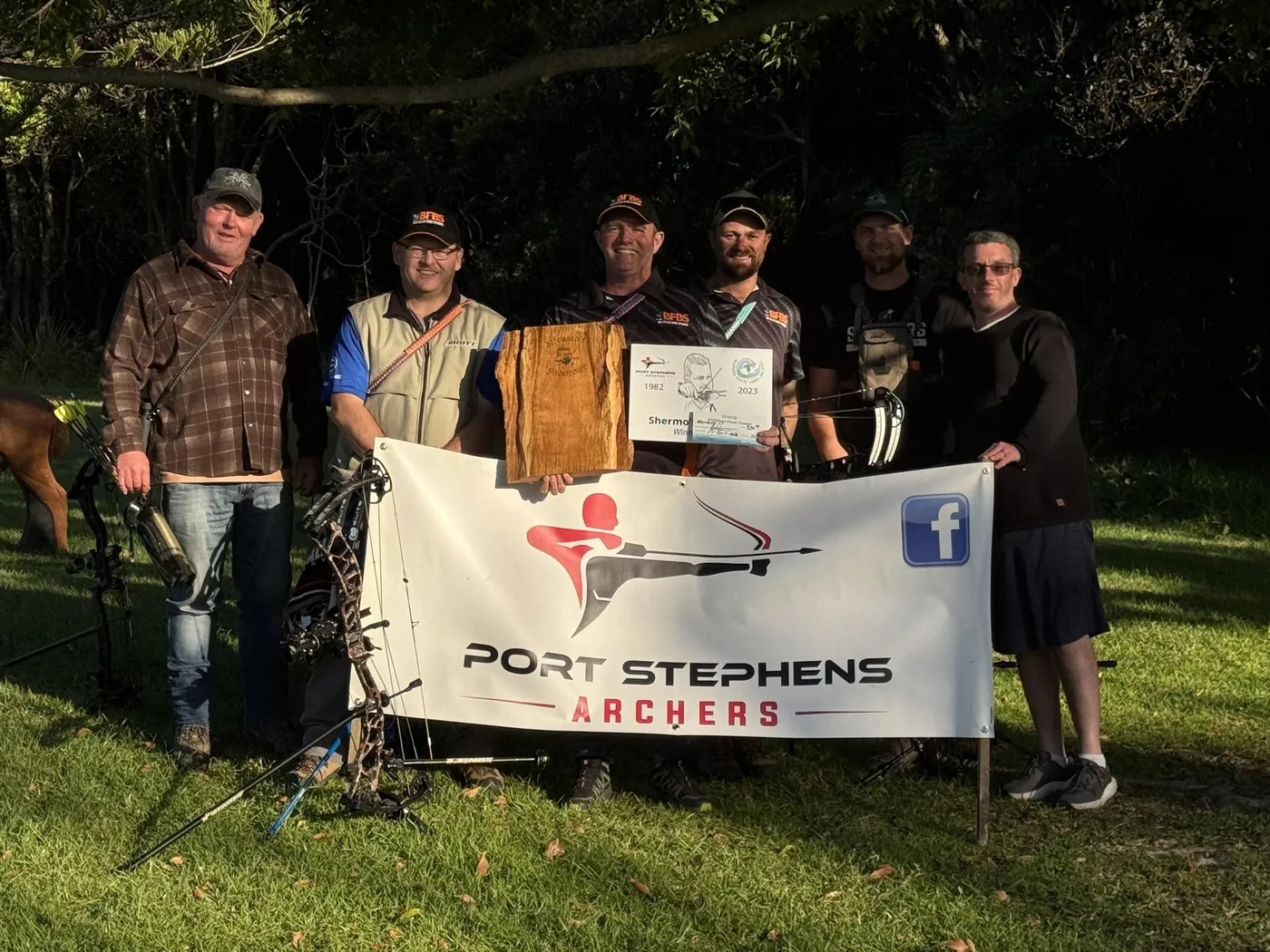 A group of archers behind a club banner at a shoot, holding a certificate and an award.