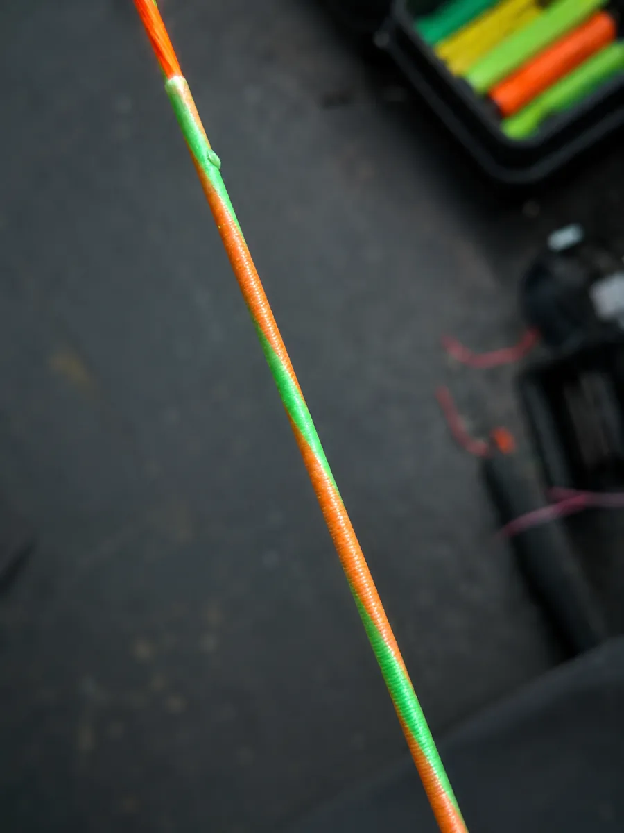 Close-up of a two-colour bowstring in orange and green with spools behind it on the bench.