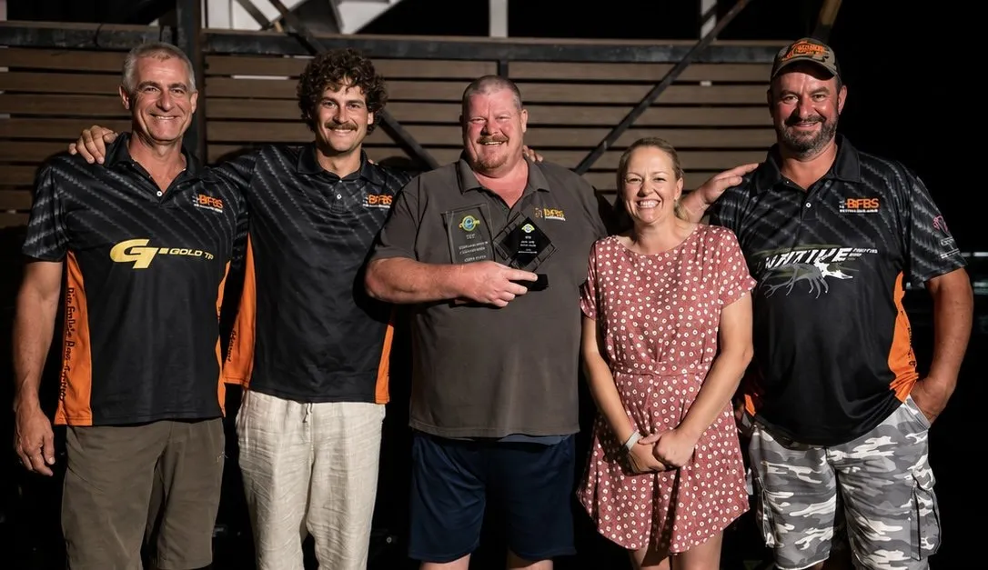 Five archers and supporters standing together in the evening, three of them in Big Fella's Bow Strings shirts, one holding a pair of trophies.