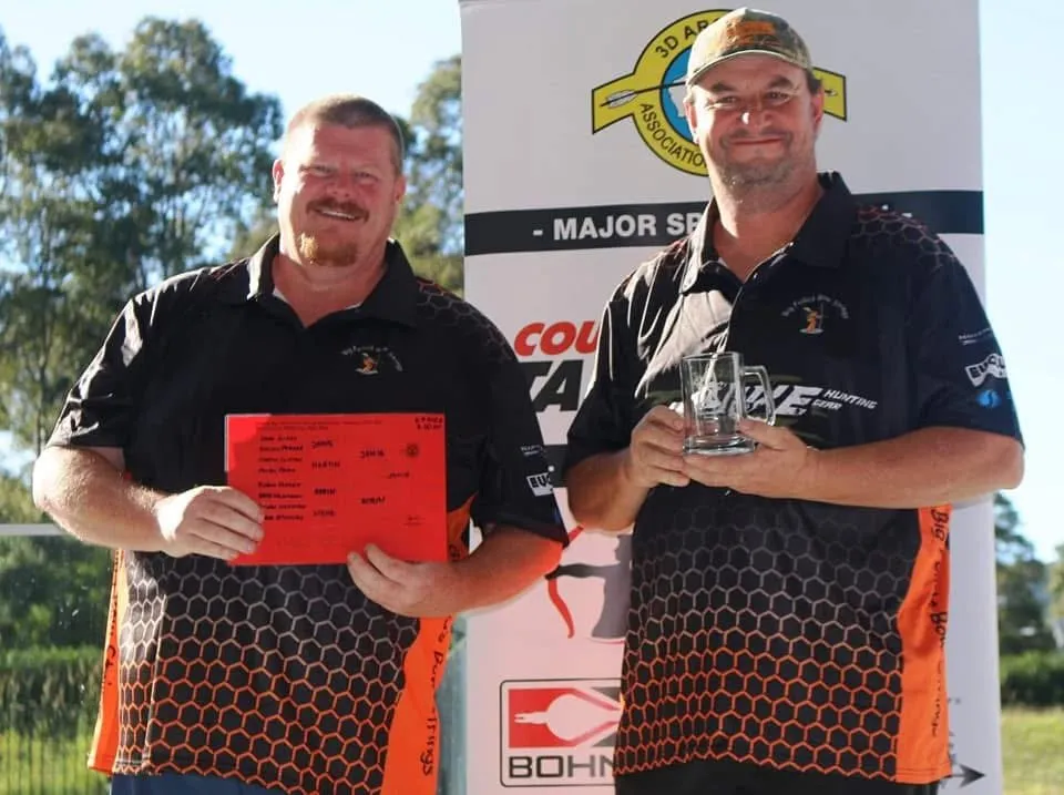 Two archers holding a certificate and a glass trophy in front of an event banner.